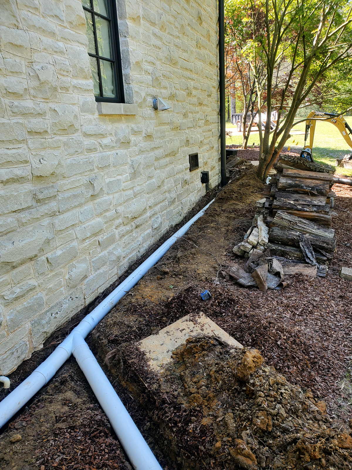 PVC drainage pipes installed along stone house foundation in trench, wood pile and mini excavator nearby